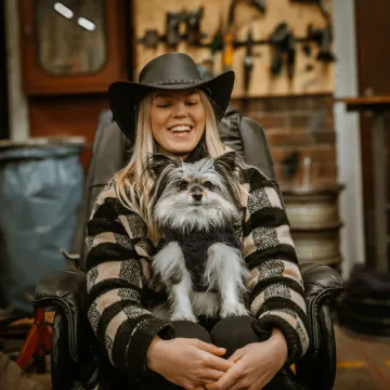 Skrattande kvinna med hatt håller en liten hund i en fåtölj