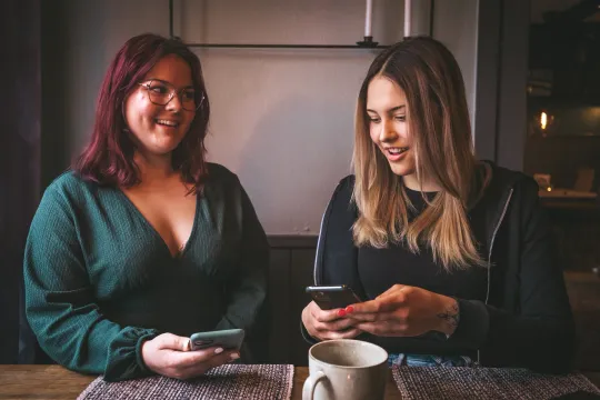 Två unga kvinnor tittar på sina mobiler vid ett bord