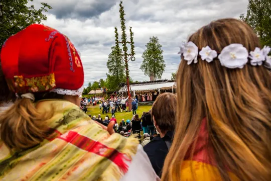 Två personer i folkdräkt och blomsterkrans framför en midsommarstång
