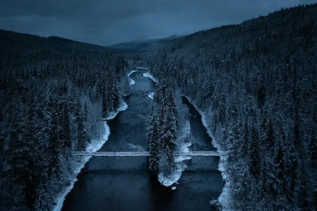 Flod med bro genom snöig granskog sedd från ovan i skymning