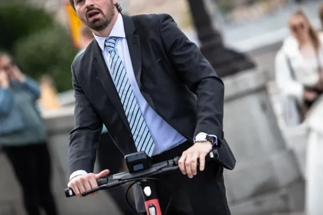Man i kostym och slips står med en elsparkcykel på en gata