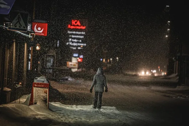 Person går längs en snöig gata med butiksskyltar i snöfall på natten