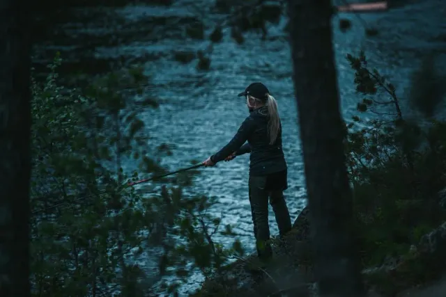 Person med keps står vid vattnet i skogen i blått kvällsljus