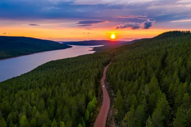 Grusväg genom skog ner mot en sjö vid solnedgång sedd från ovan