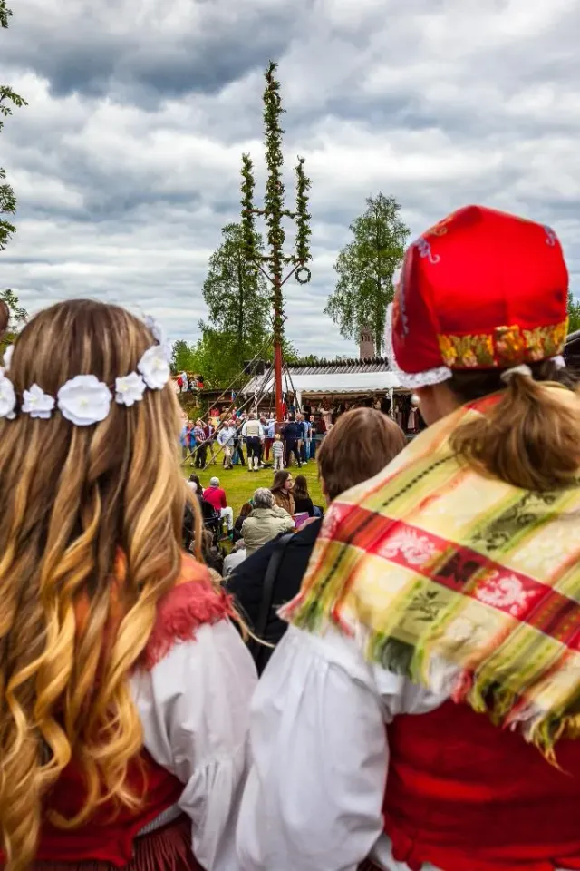 Två personer i folkdräkt och blomsterkrans framför en midsommarstång