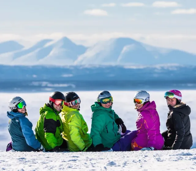 Sex personer i skidkläder och hjälmar sitter i snön med fjäll bakom