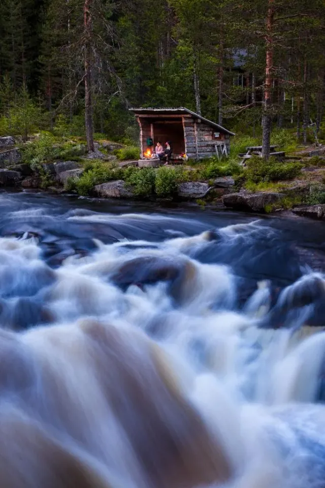 Strömmande fors med liten stuga vid skogskanten