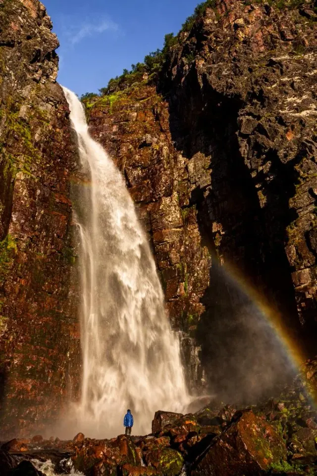 Högt vattenfall mellan klippväggar med regnbåge och en person nedanför