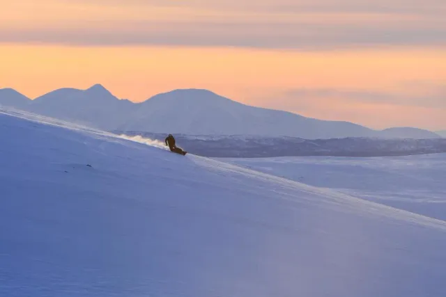 Skidåkare nedför en snöbacke mot fjäll i rosa kvällsljus