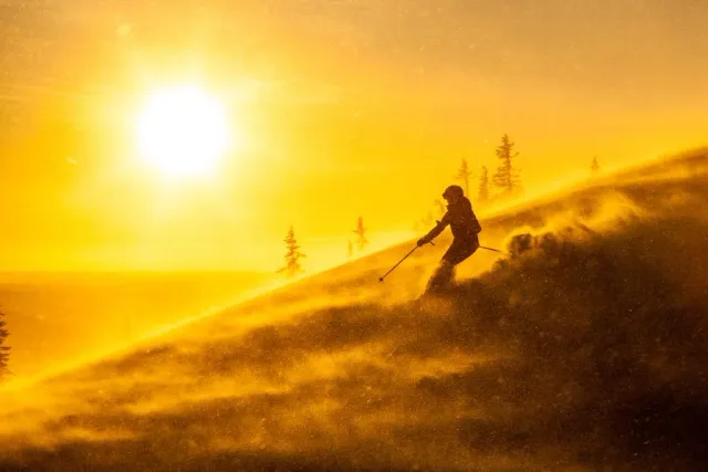 Skidåkare i motljus och snövirvlar vid låg sol