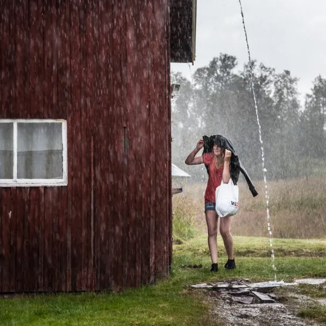 Person skyddar sig mot regn och vattenstråle vid ett rött trähus