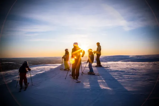 Skidåkare står på en snöig höjd mot låg sol