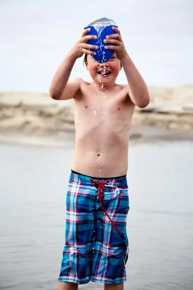 Pojke häller vatten över huvudet med en blå hink på en strand