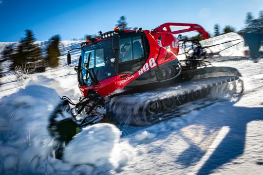Röd pistmaskin kör genom snö i solsken