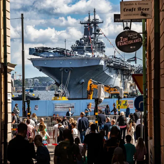 Stort örlogsfartyg i hamn sett från en trång gata med folk