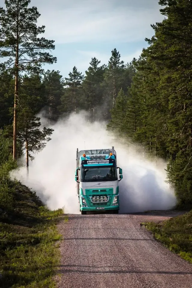 Lastbil kör på grusväg genom skog med stort dammoln