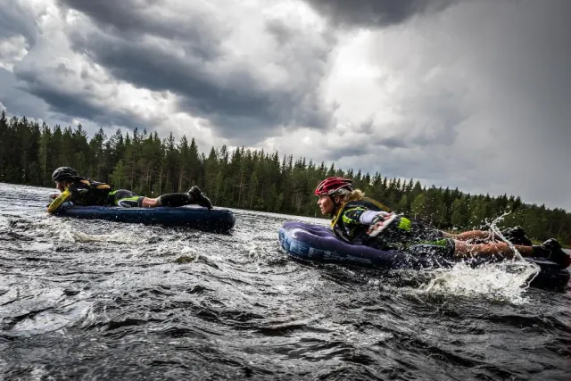Två personer paddlar på luftmadrasser i en älv under mörka moln