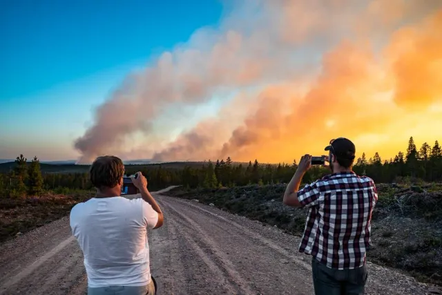 Två personer fotograferar en stor rökpelare vid en grusväg i solnedgång