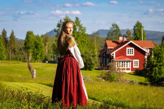 Kvinna i folkdräkt står på en äng framför ett rött hus