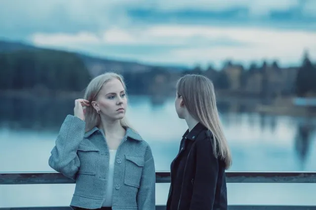 Två unga kvinnor står vid ett räcke framför en sjö