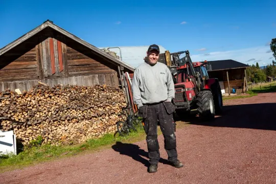 Man står framför en traktor och en vedtrave vid en lada