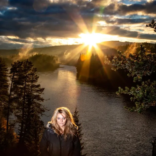 Kvinna står vid en älv med solen bakom skogen