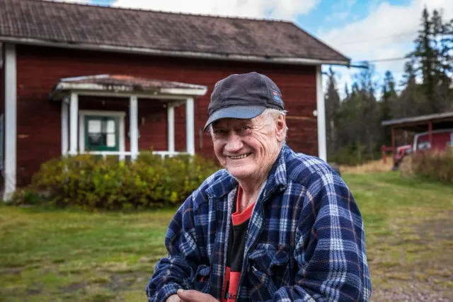 Leende man med keps och rutig skjorta framför ett rött hus
